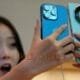 Wanita memegang iPhone 15 dan ponsel lain dengan ekspresi terkejut melihat perbandingan harga