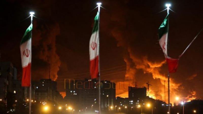 Bendera Iran berkibar saat api dan asap dari serangan Israel terhadap depot minyak Sharan membubung, menyusul serangan Israel terhadap Iran, di Teheran, Iran, 15 Juni 2025. (via REUTERS/Majid Asgaripour)